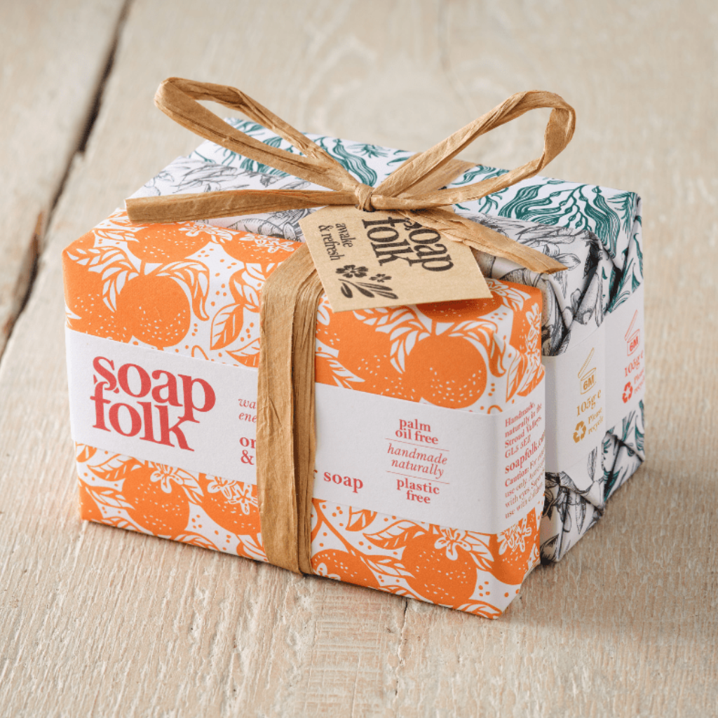 Three bars of Soap Folk soap wrapped in colourful botanical patterned paper, tied with a brown raffia ribbon and brown paper tag, placed on a light wood surface. The front soap is Orange &amp; Ginger, the middle soap is Lemon &amp; Bergamot, and the final soap is Rosemary &amp; Kelp.