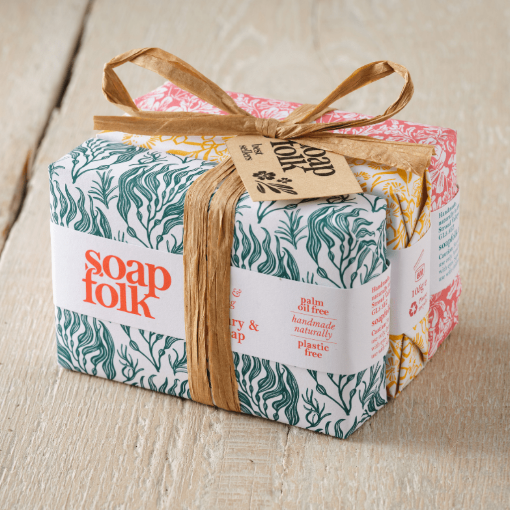 Three bars of Soap Folk soap wrapped in colourful botanical patterned paper, tied with a brown raffia ribbon and brown paper tag, placed on a light wood surface. The front soap is Rosemary & Kelp, the middle soap is Goats' Milk and Honey, and the final soap is Rose Geranium.