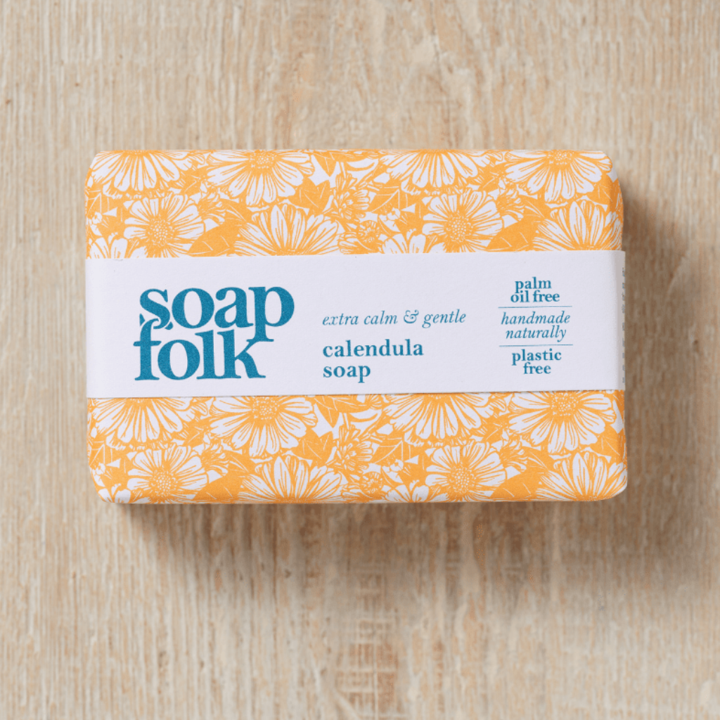 Bar of 'soapfolk' Calendula soap with yellow calendula flower packaging. Blue text states: 'soapfolk, extra calm & gentle, calendula soap, palm oil free, handmade naturally, plastic free.' The bar is placed on a light wooden surface. 