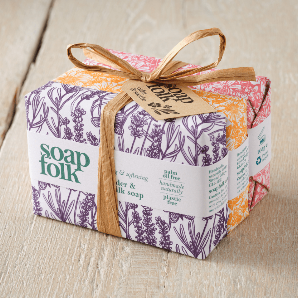 Three bars of Soap Folk soap wrapped in colourful botanical patterned paper, tied with a brown raffia ribbon and brown paper tag, placed on a light wood surface. The front soap is Rosemary & Kelp, the middle soap is Goats' Milk and Honey, and the final soap is Rose Geranium.