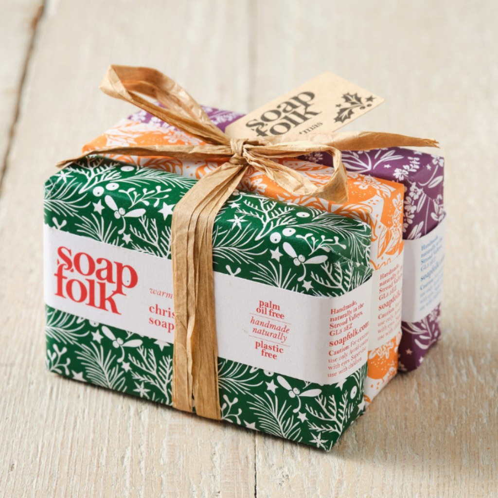 Three wrapped bars of Soap Folk Christmas soap gift set on a light wood background. The front bar is christmas soap wrapped in green pattern paper with a white label, and the two bars behind it show orange - orange and ginger soap -and purple - night lavender soap - patterned wrapping tied with a brown raffia ribbon.