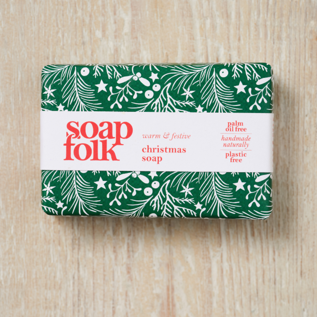 A wrapped bar of soap from the brand 'soap folk' with a green and white Christmas holly pattern and red text stating: 'soapfolk, warm &amp; festive christmas soap, palm oil free, handmade naturally, plastic free'. The bar is placed on a light wood background.