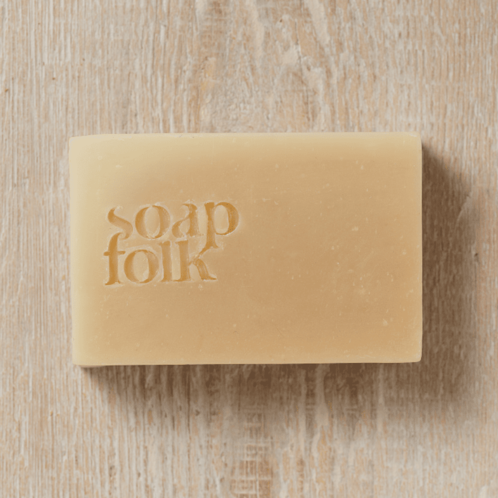 Bar of Goats' Milk and Honey soap with packaging depicting yellow honeycomb and bees with red text stating: 'soapfolk, creamy & gentle, palm oil free, handmade naturally, plastic free.' Sat on a wooden surface.