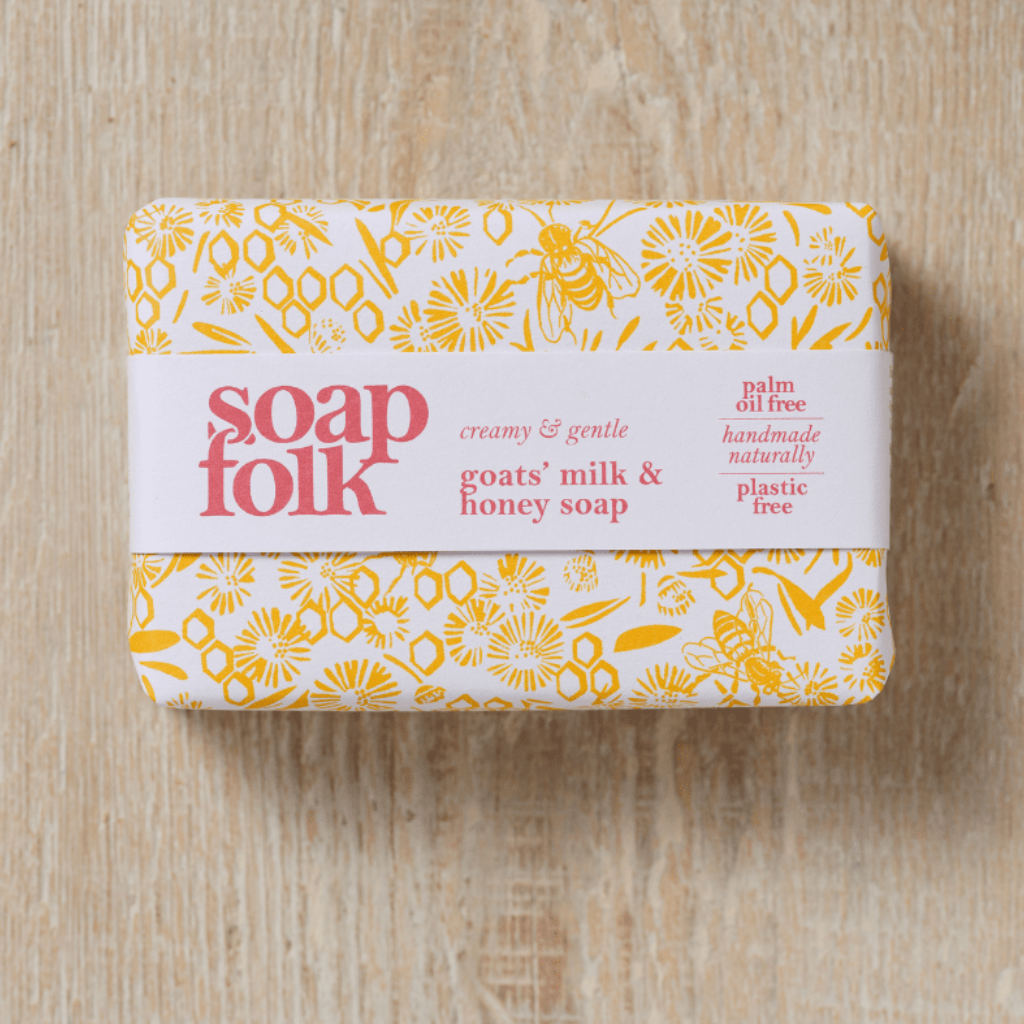 Bar of Goats' Milk and Honey soap with packaging depicting yellow honeycomb and bees with red text stating: 'soapfolk, creamy &amp; gentle, palm oil free, handmade naturally, plastic free.' Sat on a wooden surface.