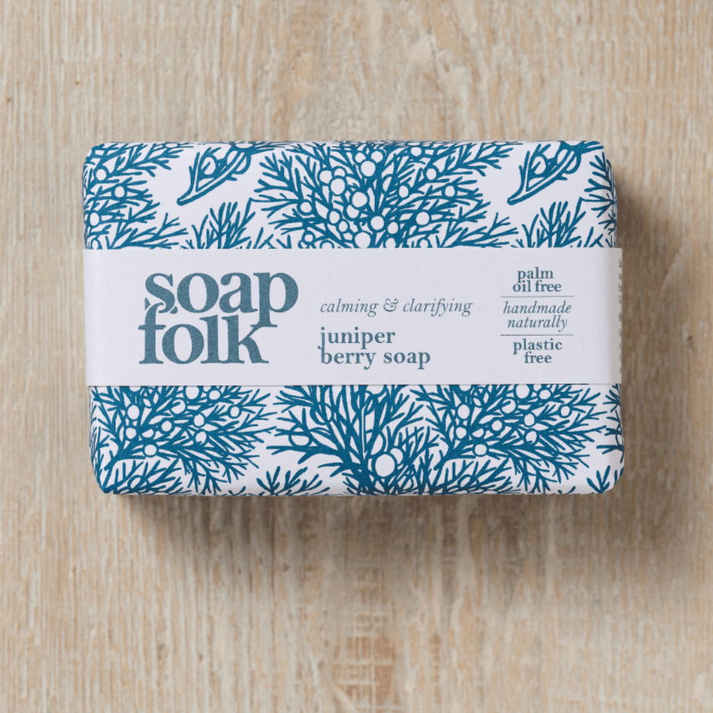 Bar of Juniper Berry soap with blue juniper berry bush illustrations. Blue text states: 'soapfolk, calming &amp; clarifying, juniper berry soap, palm oil free, handmade naturally, plastic free.' The bar is sat on a light wooden surface.