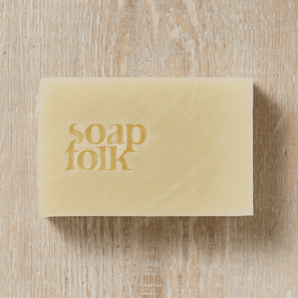 A rectangular bar of Soap Folk lavender and oatmeal soap wrapped in a white paper band with purple botanical illustrations. Teal text reads: 'soapfolk, relaxing & softening, lavender & oatmilk soap, palm oil free, handmade naturally, plastic free.' Placed on a light wood surface.
