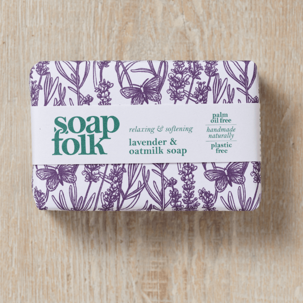A rectangular bar of Soap Folk lavender and oatmeal soap wrapped in a white paper band with purple botanical illustrations. Teal text reads: 'soapfolk, relaxing & softening, lavender & oatmilk soap, palm oil free, handmade naturally, plastic free.' Placed on a light wood surface.