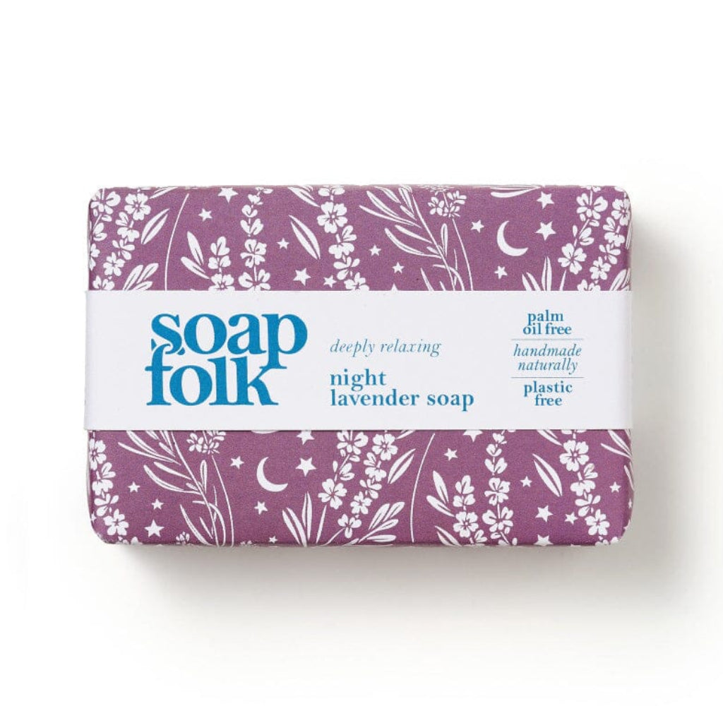 A wrapped bar of lavender soap from the brand 'soap folk'. The soap is wrapped in a purple paper sleeve decorated with white botanical illustrations of flowers, leaves, and crescent moons, and wrapped in a white paper band with blue text stating: soapfolk, deeply relaxing, night lavender soap, palm oil free, handmade naturally, plastic free.' The bar sits on a light wood background.