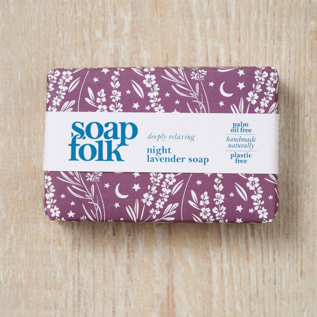 A wrapped bar of lavender soap from the brand 'soap folk'. The soap is wrapped in a purple paper sleeve decorated with white botanical illustrations of flowers, leaves, and crescent moons, and wrapped in a white paper band with blue text stating: soapfolk, deeply relaxing, night lavender soap, palm oil free, handmade naturally, plastic free.' The bar sits on a light wood background.