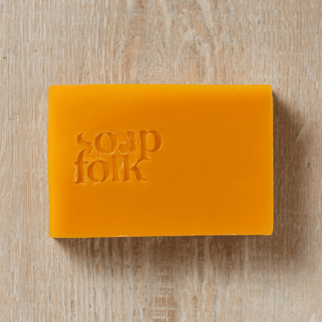 A rectangular bar of vibrant orange soap with the brand name 'soap folk' embossed on the front, resting on a light beige wood grain surface.