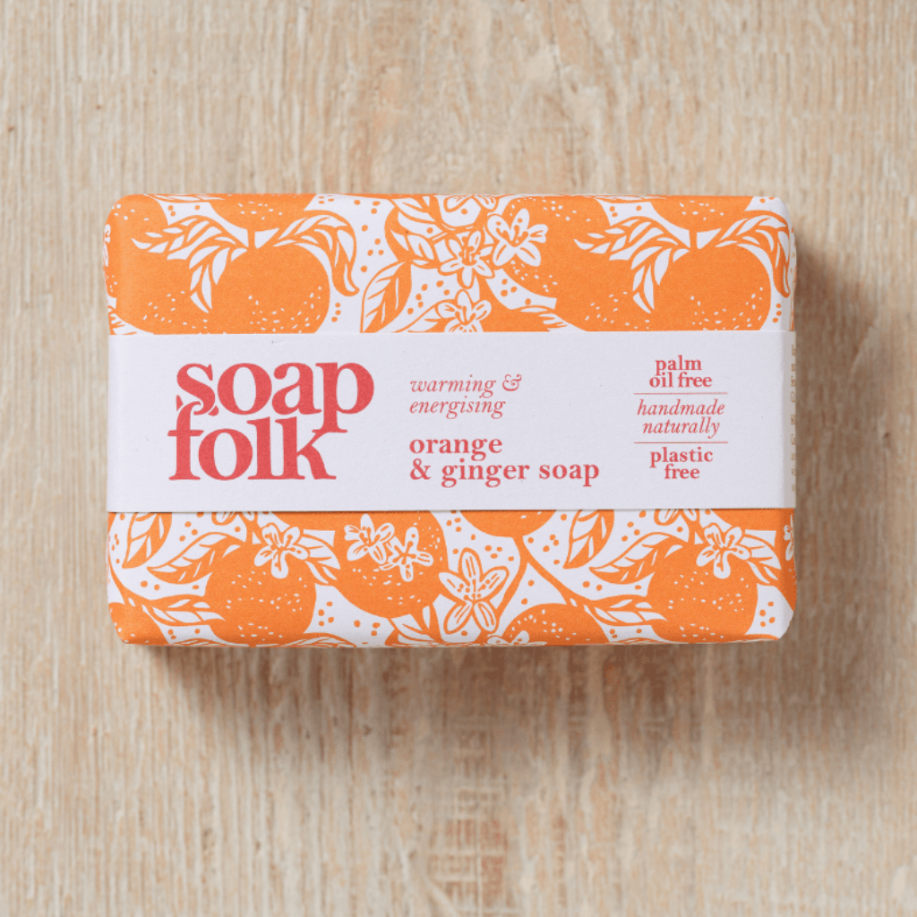Three wrapped bars of Soap Folk Christmas soap gift set on a light wood background. The front bar is christmas soap wrapped in green pattern paper with a white label, and the two bars behind it show orange - orange and ginger soap -and purple - night lavender soap - patterned wrapping tied with a brown raffia ribbon.
