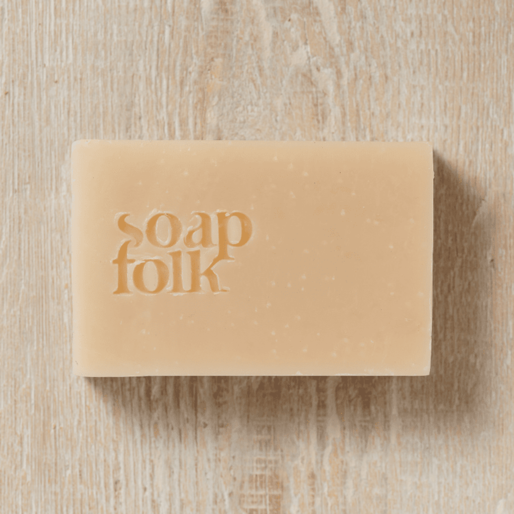 Bar of soap with a pink floral packaging design and blue text stating: 'soapfolk, sweet & soothing, rose geranium soap, palm oil free, handmade naturally, plastic free.' Placed on a wooden surface.