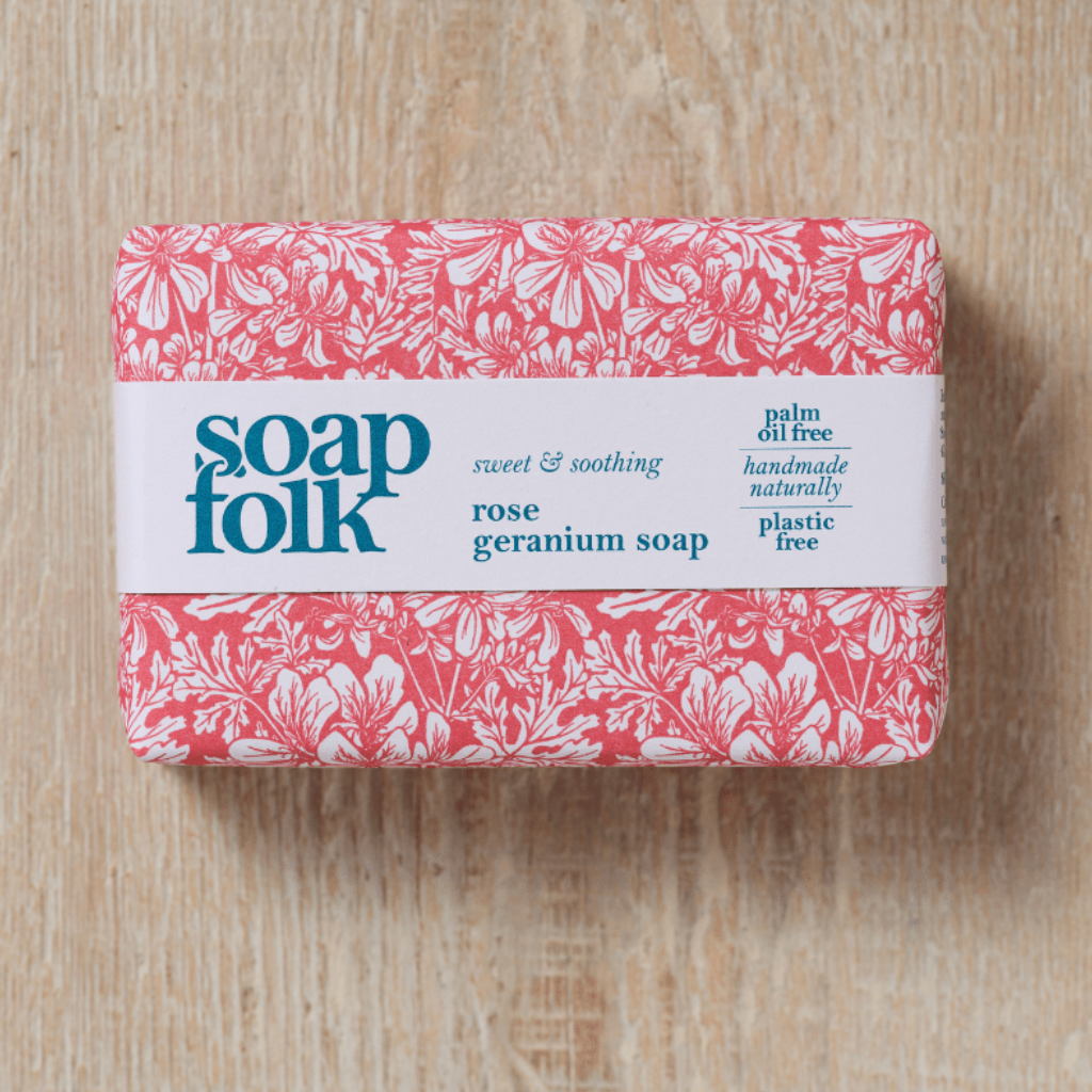Bar of soap with a pink floral packaging design and blue text stating: 'soapfolk, sweet & soothing, rose geranium soap, palm oil free, handmade naturally, plastic free.' Placed on a wooden surface.