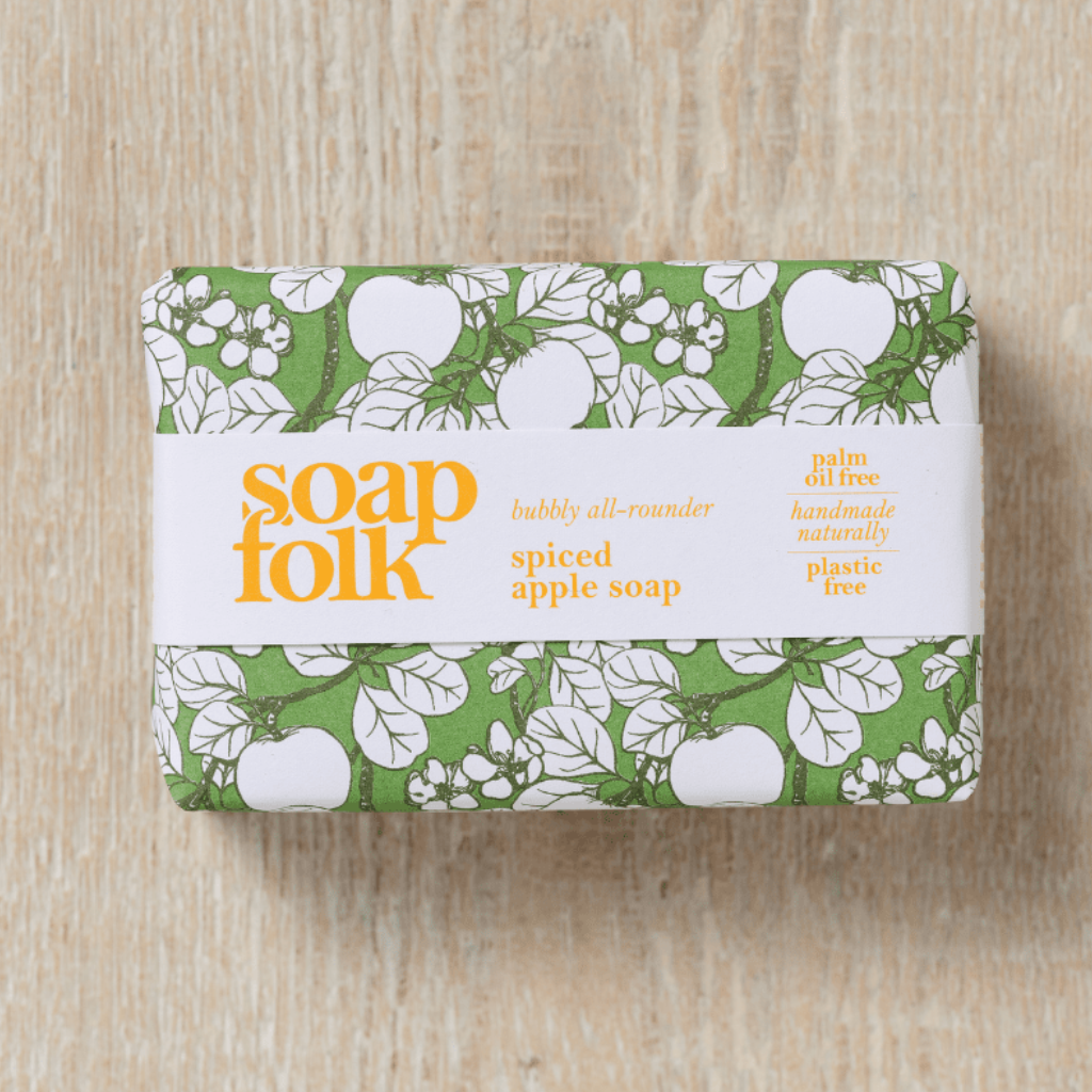 Bar of 'soapfolk' Spiced Apple soap with green apple packaging and orange/yellow text stating: 'soapfolk, bubbly all-rounder, spiced apple soap, palm oil free, handmade naturally, plastic free.' The bar is placed on a light wooden surface. 